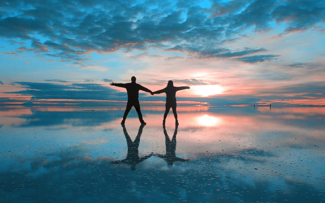 Two people holding hands on a wet beach, with the sun setting and visible below clouds
