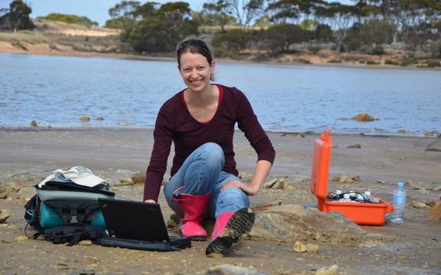 Bethany Elhmann with a laptop and equipment, working on the beach of a lake