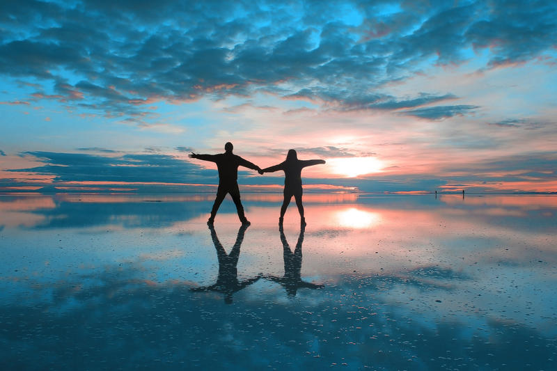 Two people holding hands on a wet beach, with the sun setting and visible below clouds