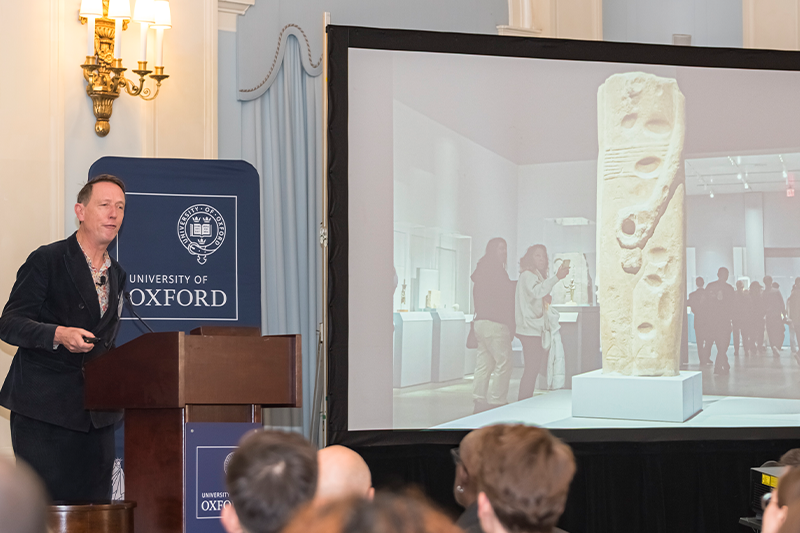 Dr Xa Sturgis speaks standing at a podium beside a large screen displaying an image of an ancient statue of the god Min