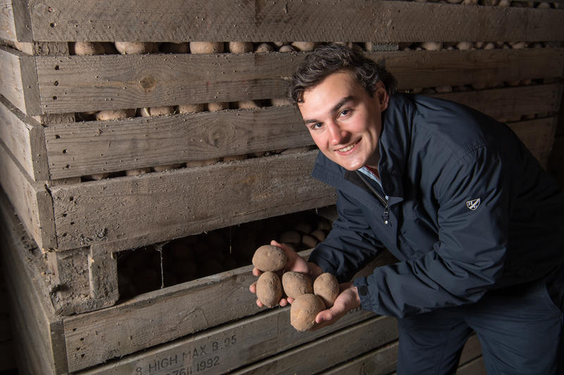 Josh Burton holding potatoes, stood next to large storage crates of potatoes