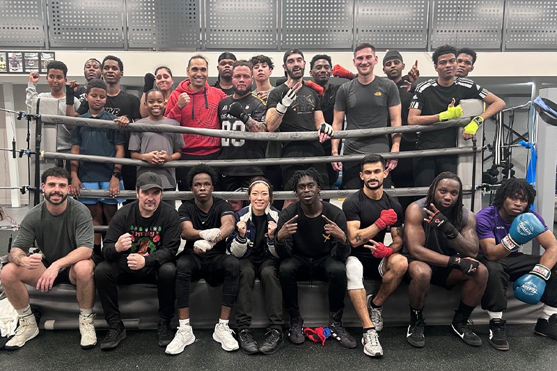 Julia Lee sits at the front of a large group of boxers in a gym, posing together inside a boxing ring at Rebels Boxing Gym