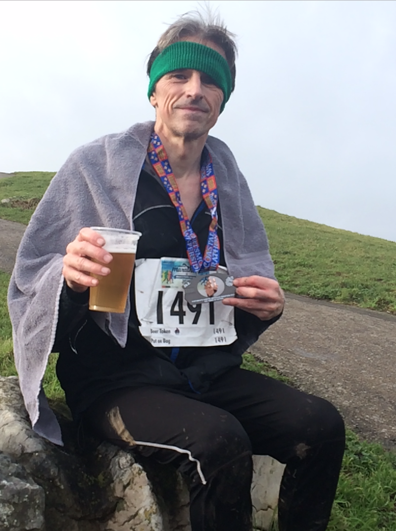 Phil, wearing running kit and a race number, holding a beer and displaying his medal for the camera, whilst sat on a large stone with the sea in the distance