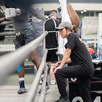 Julia Lee at ringside in a boxing gym, watching and coaching a boxer during training