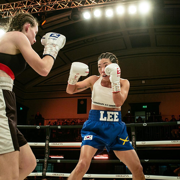 Julia Lee competes in a boxing match, wearing blue shorts and gloves, facing an opponent inside the ring under bright lights.